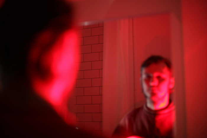 A man looks at his reflection in a mirror, bathed in eerie red light, encountering creepy and mysterious things.