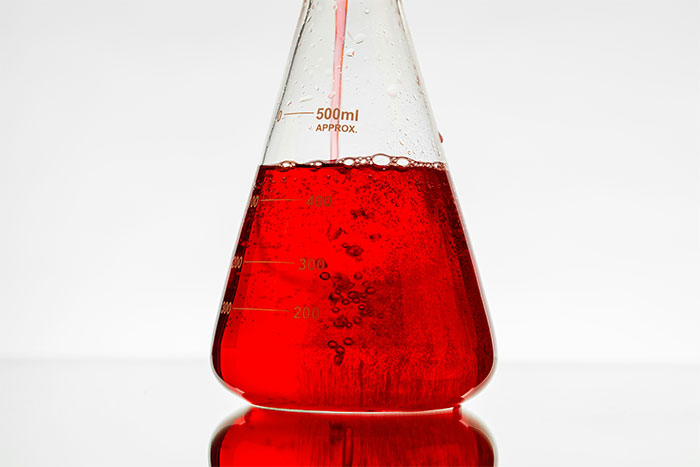 A close-up shot of a clear glass Erlenmeyer flask filled with a bubbling red potion, reflecting on a white surface.