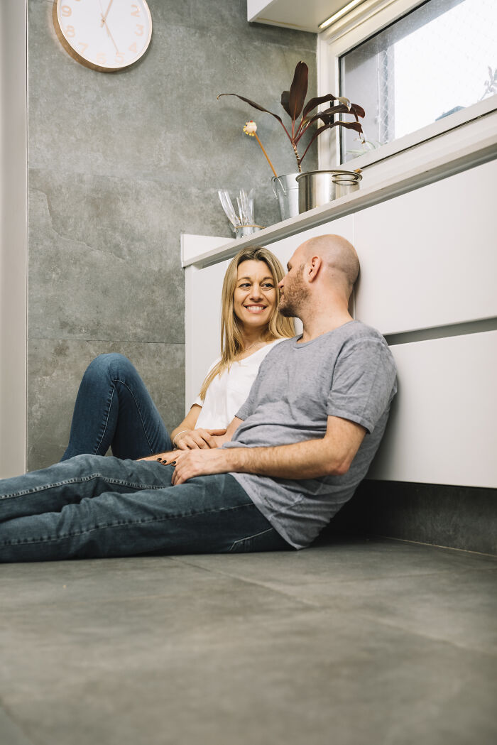 A happy age-gap couple sitting on a kitchen floor. They are smiling at each other.