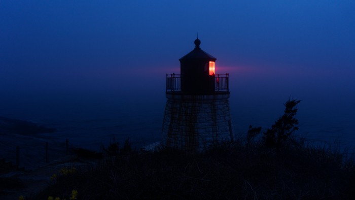A mysterious lighthouse with a glowing red light at dusk, shrouded in blue fog, representing mysterious things.