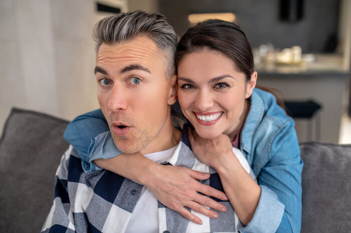 A man with a surprised expression and a smiling woman hugging him from behind, showcasing an age-gap relationship.