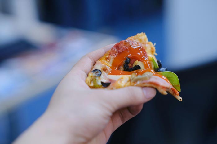 A hand holding a slice of pizza covered in an excessive amount of sauce, an unhygienic food preparation example.