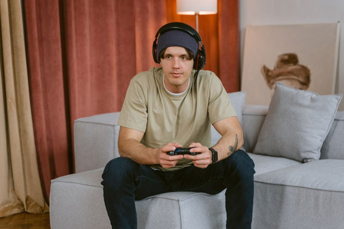 A man wearing headphones and a beanie plays video games on a couch with a controller, experiencing MIL trying to evict DIL.