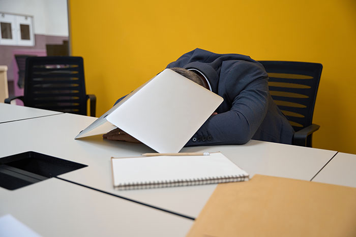 A software engineer, tired of a hiring scam, has his head on a desk, covered by a laptop in an office.