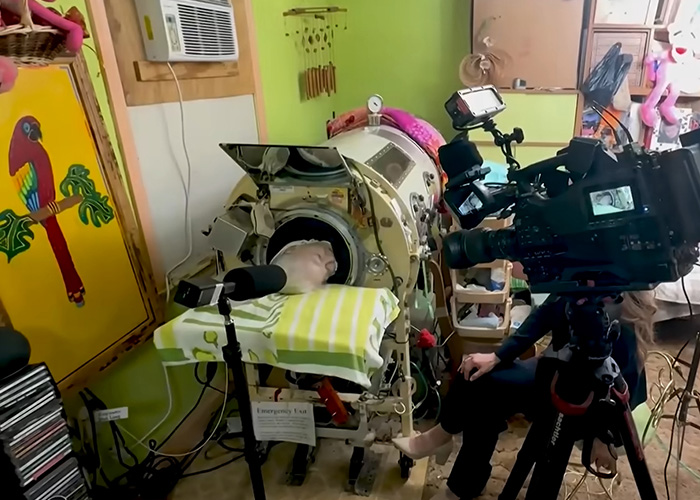 An empty iron lung machine in a room, with a camera set up for filming the polio survivor's story.