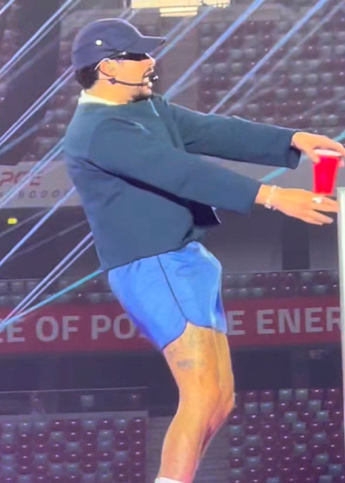 Bad Bunny performing a steamy dance move on stage in blue shorts, holding a red cup, fans lose it. Bad Bunny performing a steamy dance move on stage in blue shorts, holding a red cup, fans lose it.