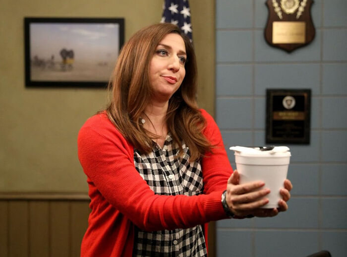 A female celebrity in a red cardigan and plaid shirt, holding a white cup, hinting at disillusioned celebrities.