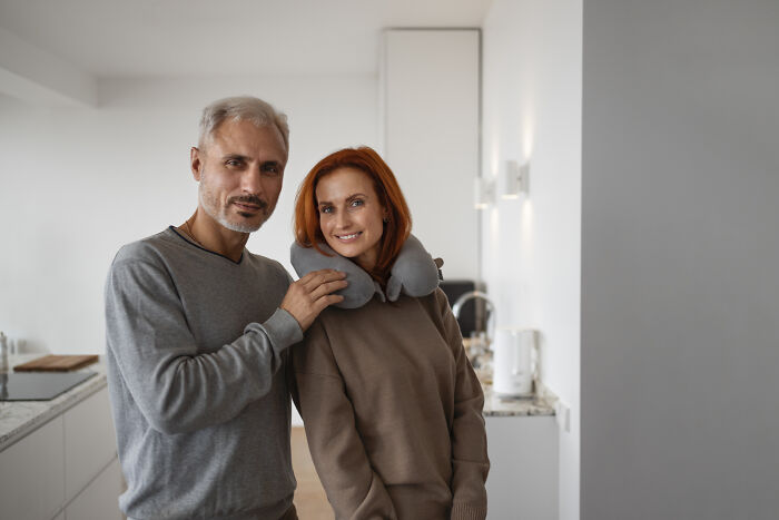 A man with grey hair and a woman with red hair, wearing a neck pillow, smiling; they are in an age-gap relationship.