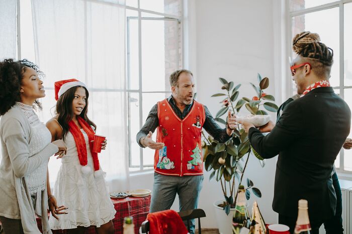 People at a party reacting to a man in an ugly Christmas sweater, potentially depicting wild stories of why people got fired.