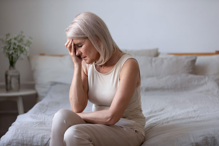 A distressed older woman with light hair sits on a bed, holding her head, reflecting the pain of harassment.