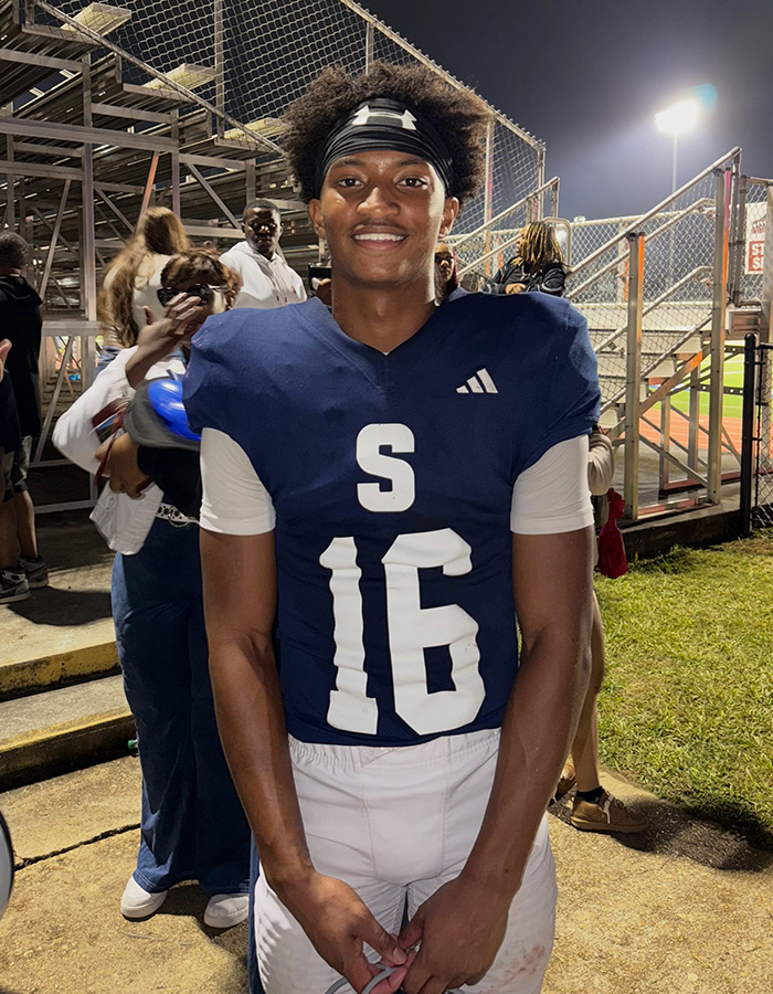 A young football player, Nolan Wells, in a blue jersey with number 16, smiles on the field.