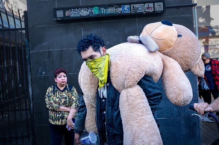 Photographer captures humor and chaos: a man with a yellow bandana carrying a giant teddy bear.
