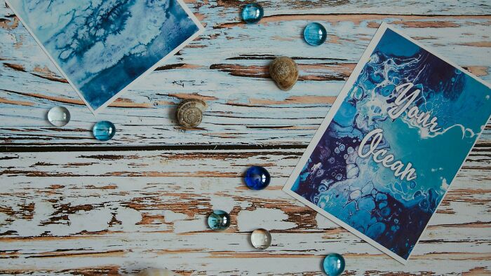 Two artistic cards with blue watercolor designs and the words Your Ocean, alongside seashells and glass beads on a wooden surface.