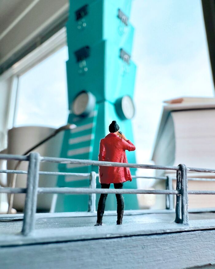 Artist Derrick Lin crafts an intricate diorama set on his office desk, featuring a figure looking up at a tall, teal structure.