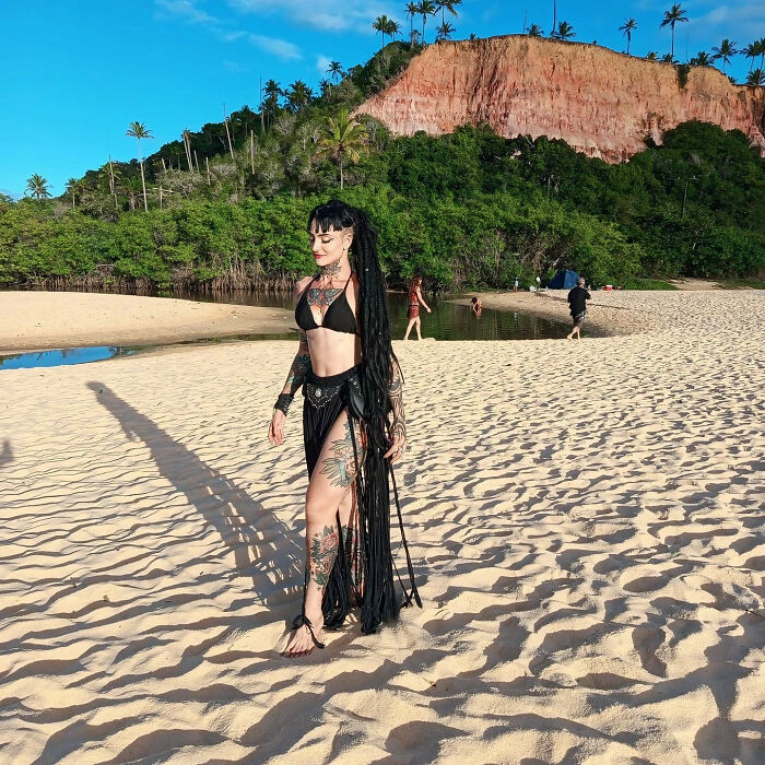 A Goth influencer with long dark dreadlocks and tattoos walks on a sandy beach next to a lagoon. A Goth influencer with long dark dreadlocks and tattoos walks on a sandy beach next to a lagoon.