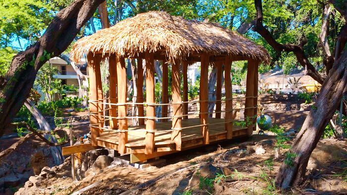 A newly built thatched-roof gazebo, a creative home DIY project, sits nestled among trees in a sunny outdoor setting.
