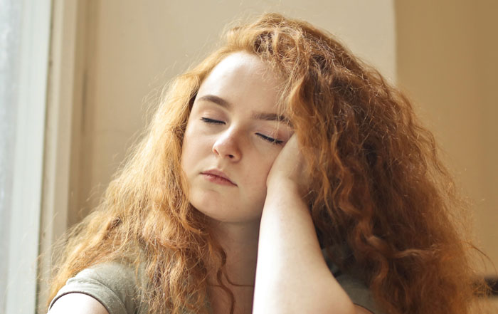 A teen with long, curly red hair rests her head on her hand, eyes closed, tired from being forced to eat. A teen with long, curly red hair rests her head on her hand, eyes closed, tired from being forced to eat.