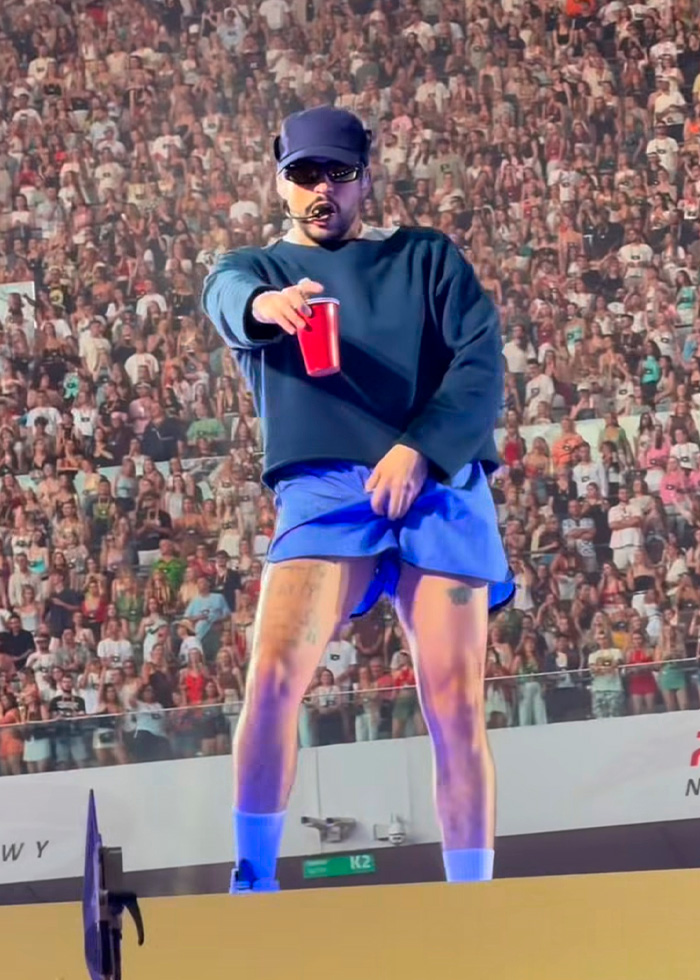 Bad Bunny in blue shorts and a dark long-sleeve shirt, holding a red cup, performing a steamy dance move on stage. Bad Bunny in blue shorts and a dark long-sleeve shirt, holding a red cup, performing a steamy dance move on stage.