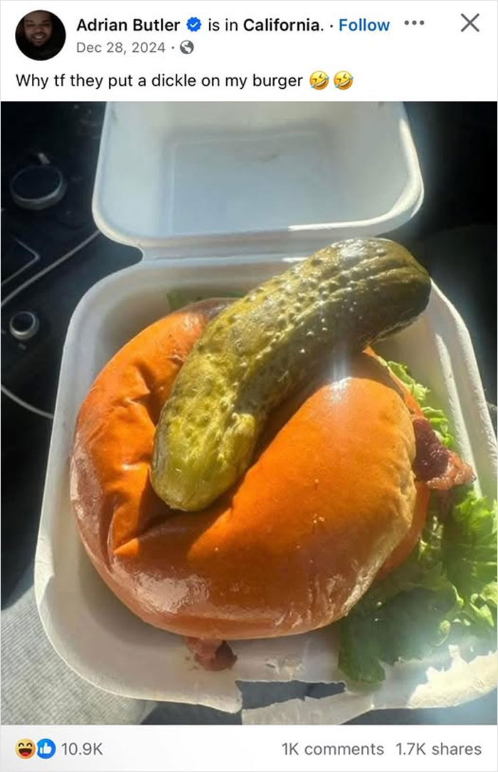 An absurd screenshot of a whole pickle on top of a burger in a take-out container.