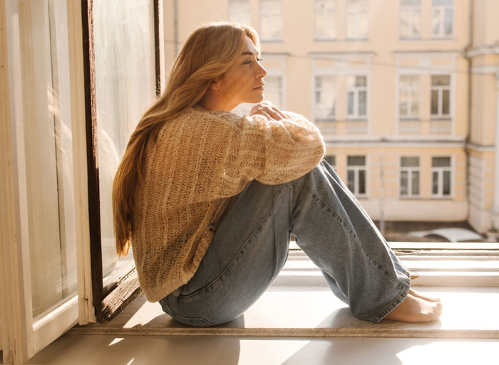 A woman sitting on a window sill, looking out contemplatively, sharing hard truths women learned.