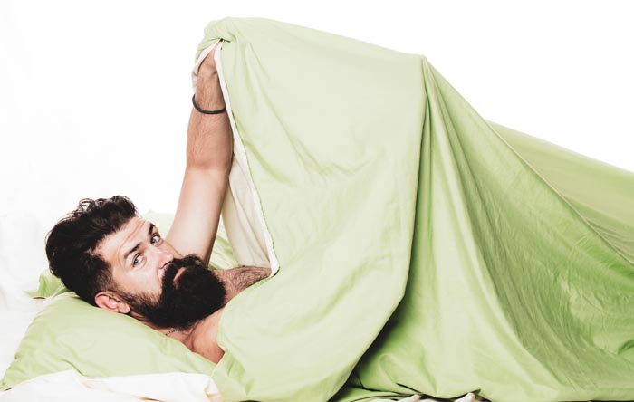 A bearded man looking from under a green duvet in bed, depicting something unhygienic.