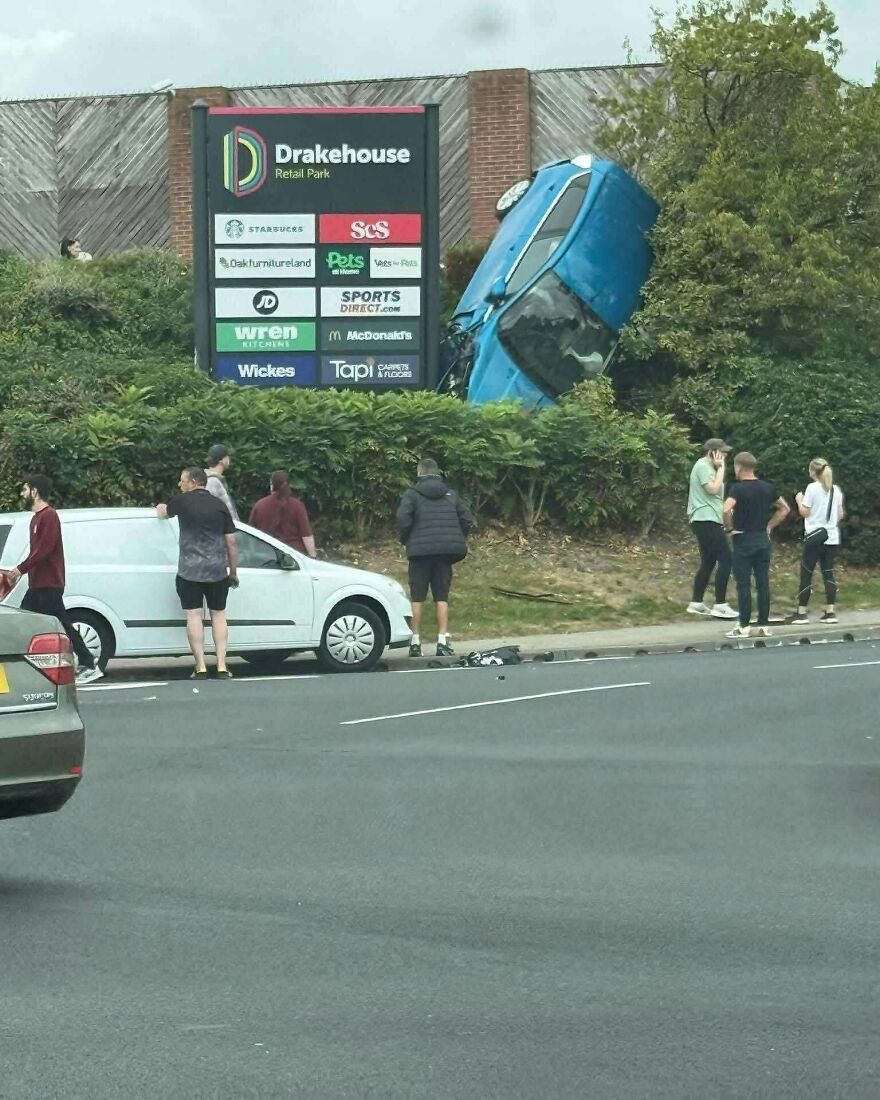 A blue car crashed into a bush next to a retail park sign, a rough day behind the wheel.