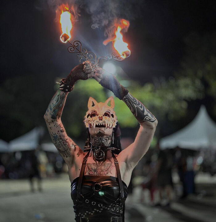 A goth influencer with a skull mask and tattoos, holding two flaming staffs during a performance. A goth influencer with a skull mask and tattoos, holding two flaming staffs during a performance.