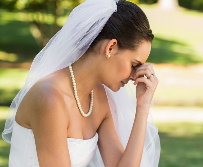 A distressed bride in a wedding dress, with a veil and pearl necklace, holds her head, symbolizing wedding stress. A distressed bride in a wedding dress, with a veil and pearl necklace, holds her head, symbolizing wedding stress.