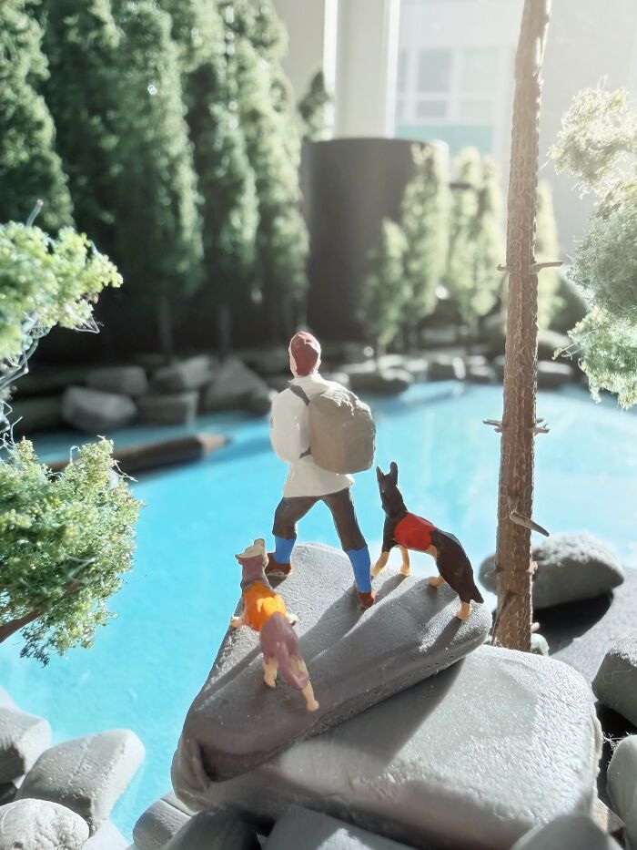 Artist Derrick Lin crafts an intricate diorama set on his office desk, featuring a hiker and dogs by a blue lake.