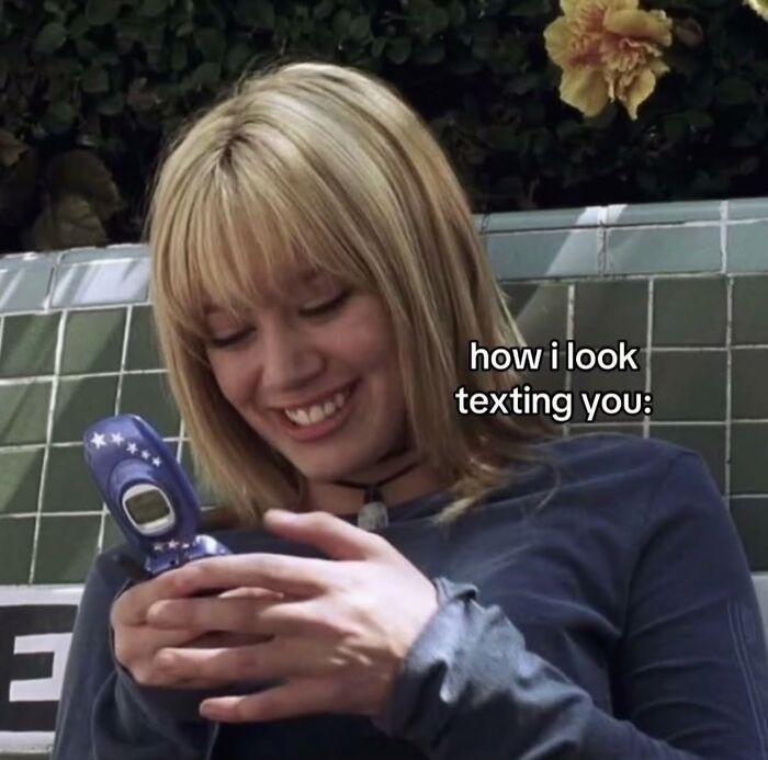 A relatable meme of a smiling woman texting on a flip phone captioned: how i look texting you.