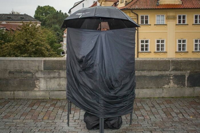 A person covered by a black tarp and umbrella on a cobblestone bridge, a serendipitous moment captured by a street photographer.