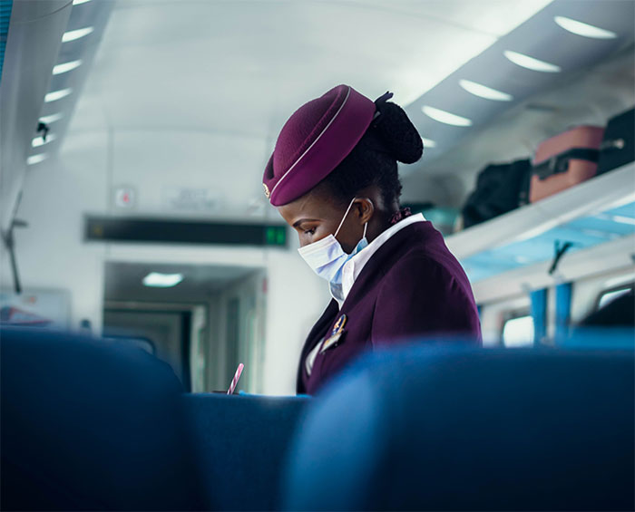 A flight attendant in uniform wearing a mask, looking down while taking notes on a flight. A flight attendant in uniform wearing a mask, looking down while taking notes on a flight.