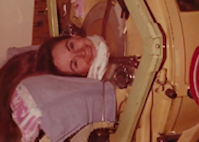 An older polio survivor woman smiles from inside her iron lung, with long hair and a white neck scarf.