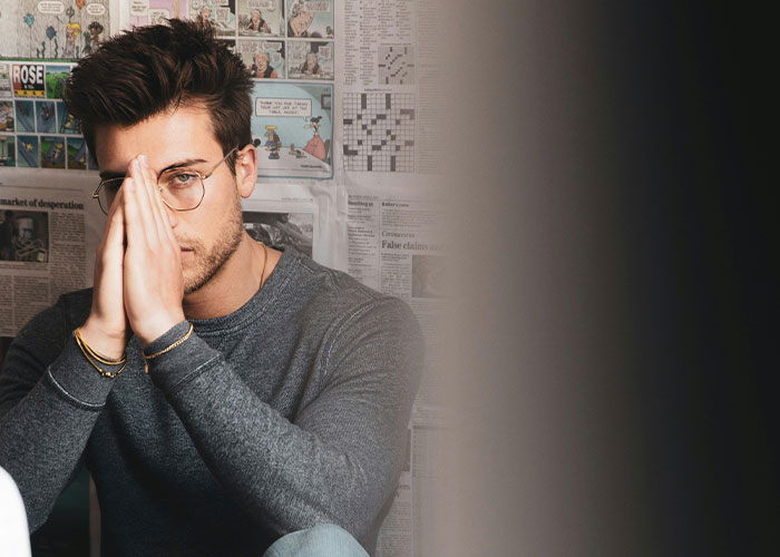 A man with glasses and a thoughtful expression, with newspapers in the background. This relates to men and their worst selves.