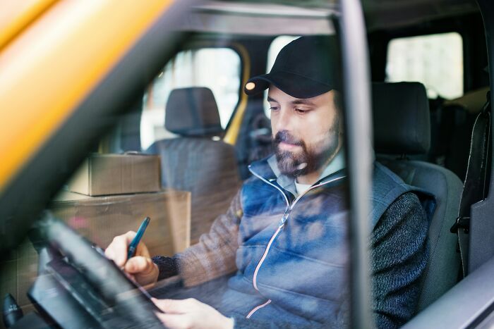 A delivery driver in a cap signing documents inside his vehicle, showing work that can lead to people getting fired from their jobs.
