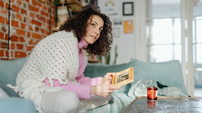 Woman in an off-white knitted shawl sitting on a turquoise couch, holding a framed family picture, pondering removing his ex.