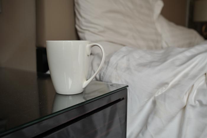 A white mug on a nightstand next to an unmade bed, indicating a new level of unhygienic behavior.