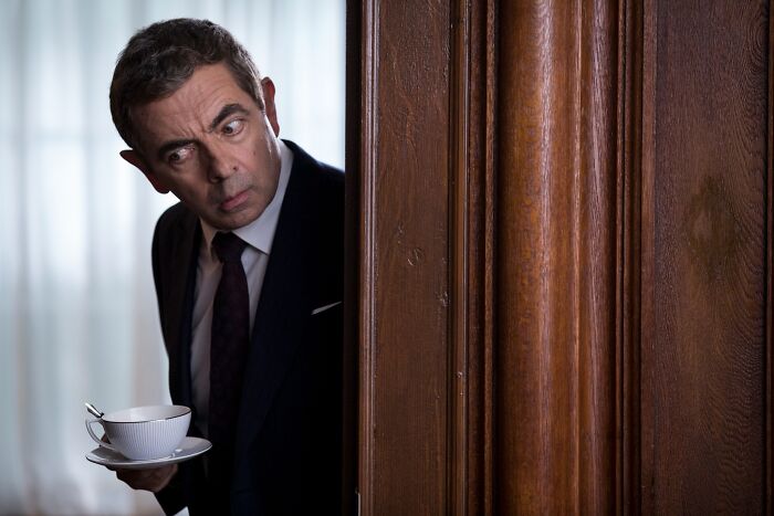 Rowan Atkinson peeking around a wooden door with a surprised expression, holding a teacup and saucer, a fascinating image.