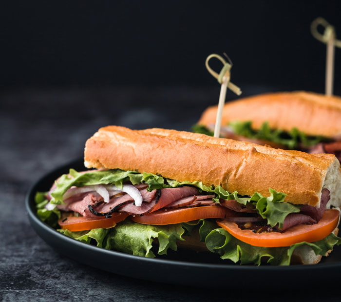 Two gourmet sandwiches with roast beef, lettuce, tomato, and onion on a dark plate, as a visual for men's complaints.