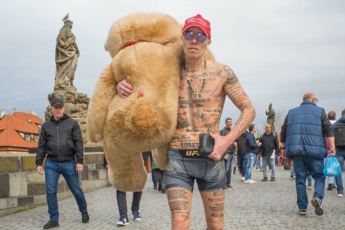 A tattooed man carrying a large teddy bear on a bridge, a serendipitous moment captured by a street photographer.