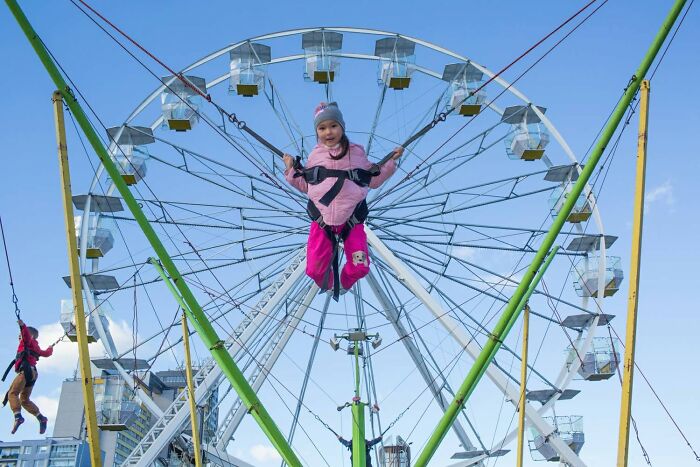 A young girl smiling while bungee jumping in front of a Ferris wheel, an everyday life moment captured by a street photographer.
