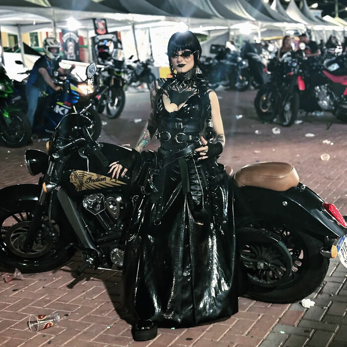 A goth influencer posing on a motorcycle, dressed in a long black vinyl dress at an outdoor event. A goth influencer posing on a motorcycle, dressed in a long black vinyl dress at an outdoor event.