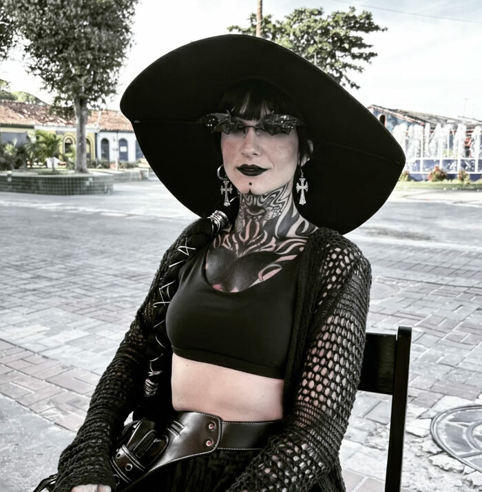 A goth influencer wearing a large black hat, sunglasses, and black outfit, sitting outdoors. A goth influencer wearing a large black hat, sunglasses, and black outfit, sitting outdoors.