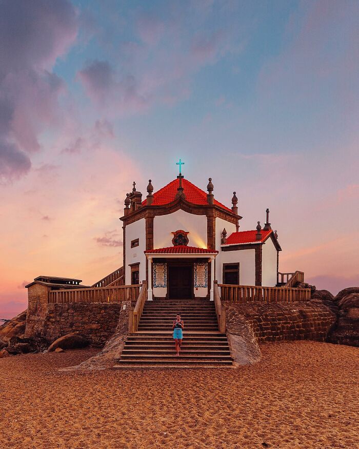 A small, white architectural location chapel with a red roof, located on a sandy beach at sunset, with a person on the stairs.