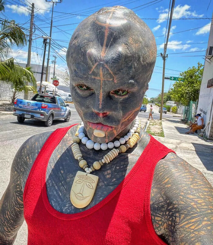 A man with extreme body modifications, known as a Black Alien, poses in a red tank top on a sunny street. A man with extreme body modifications, known as a Black Alien, poses in a red tank top on a sunny street.