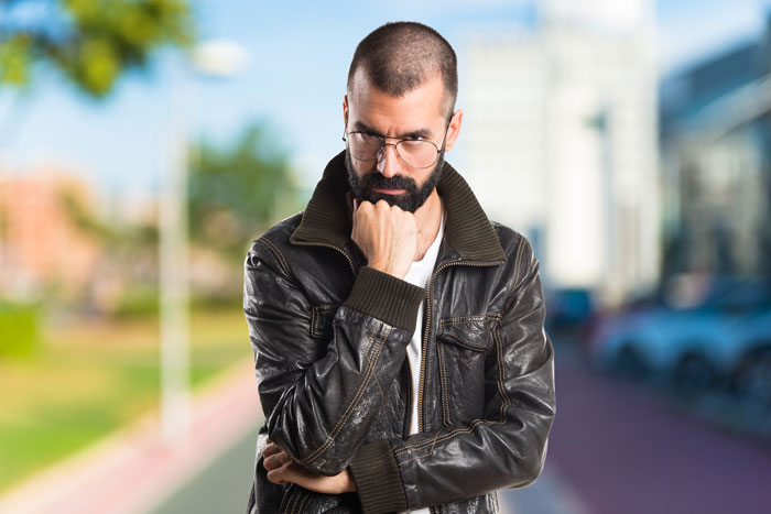 A thoughtful man with a beard and glasses, dressed in a leather jacket, appearing to ponder a missing business sign. A thoughtful man with a beard and glasses, dressed in a leather jacket, appearing to ponder a missing business sign.