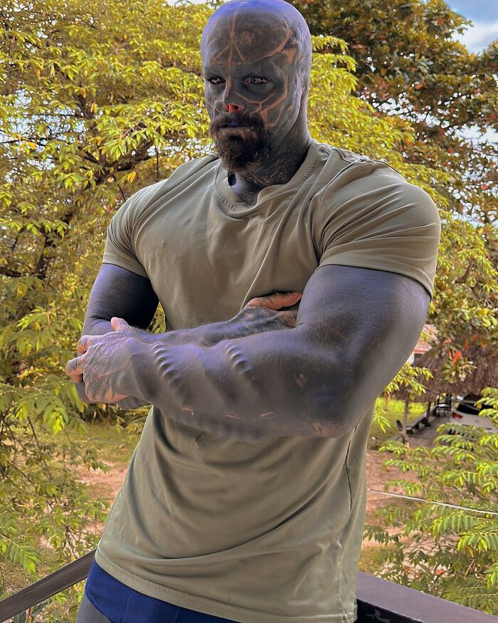 A Black Alien man with a dark beard and extensive body modifications, including textured arms, poses outdoors. A Black Alien man with a dark beard and extensive body modifications, including textured arms, poses outdoors.