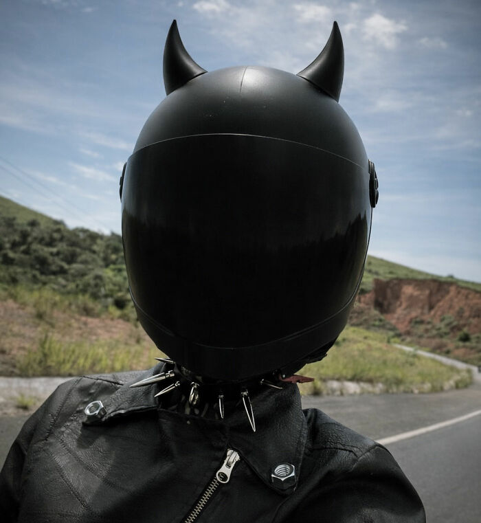 A person wearing a black motorcycle helmet with devil horns and a spiked collar on a countryside road. A person wearing a black motorcycle helmet with devil horns and a spiked collar on a countryside road.
