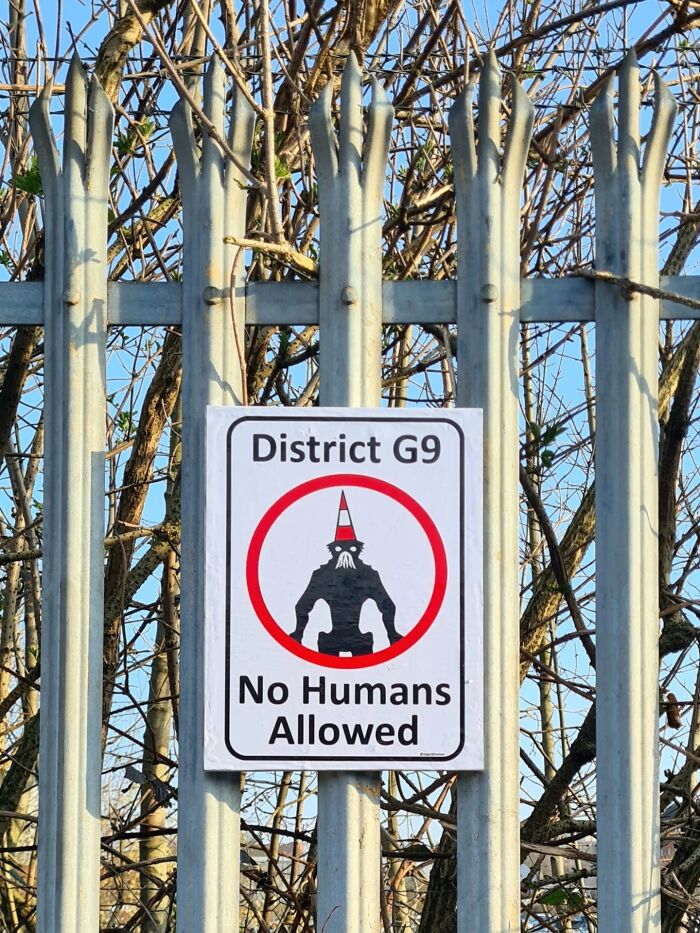 An absurd sign on a metal fence for District G9 stating No Humans Allowed with a stylized figure.
