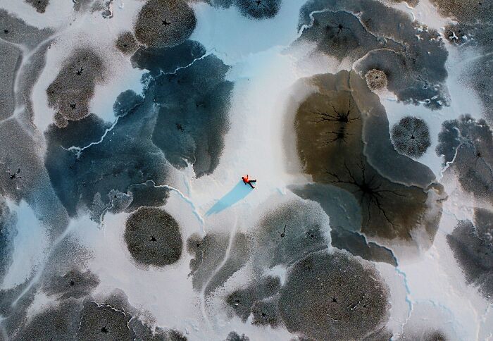 Aerial photographer captures a person lying on a vast expanse of cracked ice, surrounded by unique patterns.